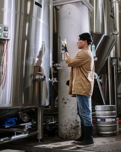 A craft brewer checking a fresh pour beside the fermenting tanks